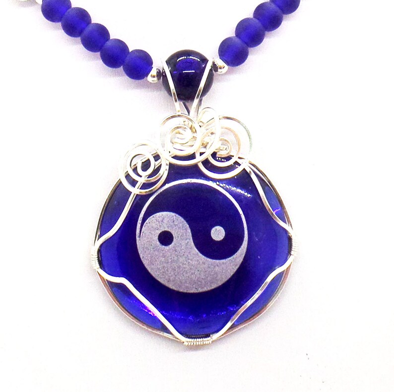 May include: A silver wire wrapped pendant necklace with a blue glass circle featuring a white yin yang symbol. The necklace is made with blue glass beads.