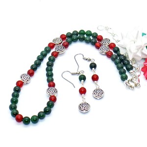 May include: A necklace and earring set featuring green and red beads with silver-toned Celtic knot accents. The necklace and earrings are displayed on a white background. The earrings have a hook closure.