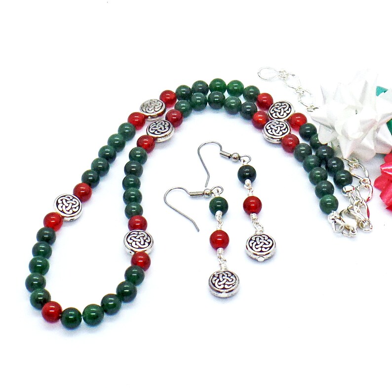 May include: A necklace and earring set featuring green and red beads with silver-toned Celtic knot accents. The necklace and earrings are displayed on a white background. The earrings have a hook closure.