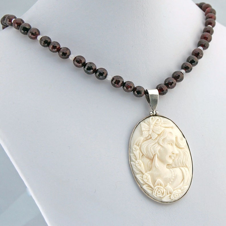 K&ouml;nnte beinhalten: Eine Halskette mit einem Anh&auml;nger, der ein geschnitztes Frauenprofil zeigt, verziert mit einem Schmetterling und Rosen. Der Anh&auml;nger h&auml;ngt an einer Kette aus dunkelroten und wei&szlig;en Perlen und bildet ein klassisches Schmuckst&uuml;ck.