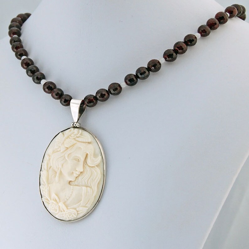 K&ouml;nnte beinhalten: Halskette mit cremefarbenem Cameo-Anh&auml;nger, der das Profil einer Frau in einem silberfarbenen Rahmen zeigt. Die Halskette hat eine Perlenkette aus dunkelroten und wei&szlig;en Perlen.