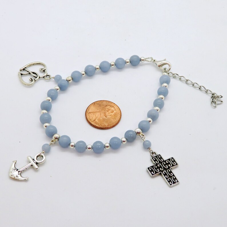 May include: A light blue beaded bracelet with silver-colored accents. The bracelet features a heart-shaped charm, an anchor charm, and a decorative cross charm. The bracelet is adjustable and has a lobster clasp closure.