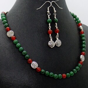 May include: A necklace and earring set featuring green and red beads. The necklace has silver-toned Celtic knot accents. The earrings have matching beads and Celtic knot charms. The jewelry is displayed on a black surface.