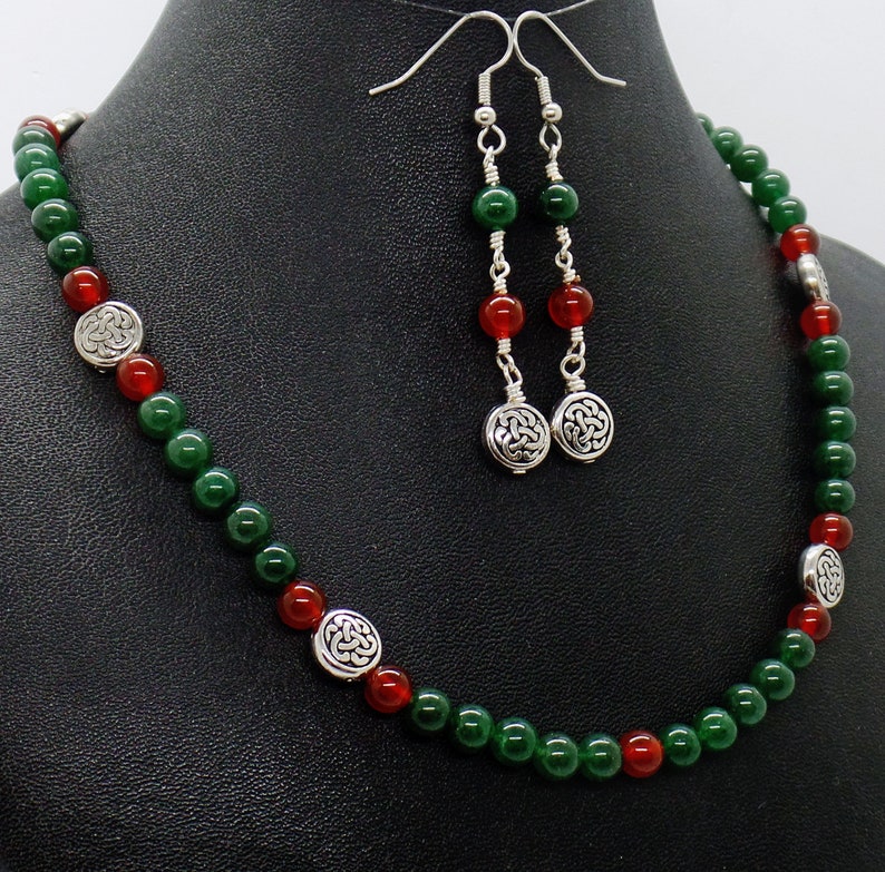 May include: A necklace and earring set featuring green and red beads. The necklace has silver-toned Celtic knot accents. The earrings have matching beads and Celtic knot charms. The jewelry is displayed on a black surface.