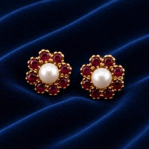 May include: A pair of gold stud earrings, each featuring a central white pearl surrounded by a cluster of eight round-cut deep red rubies. The stones are set in a floral arrangement with a polished gold metal base, creating a symmetrical design.