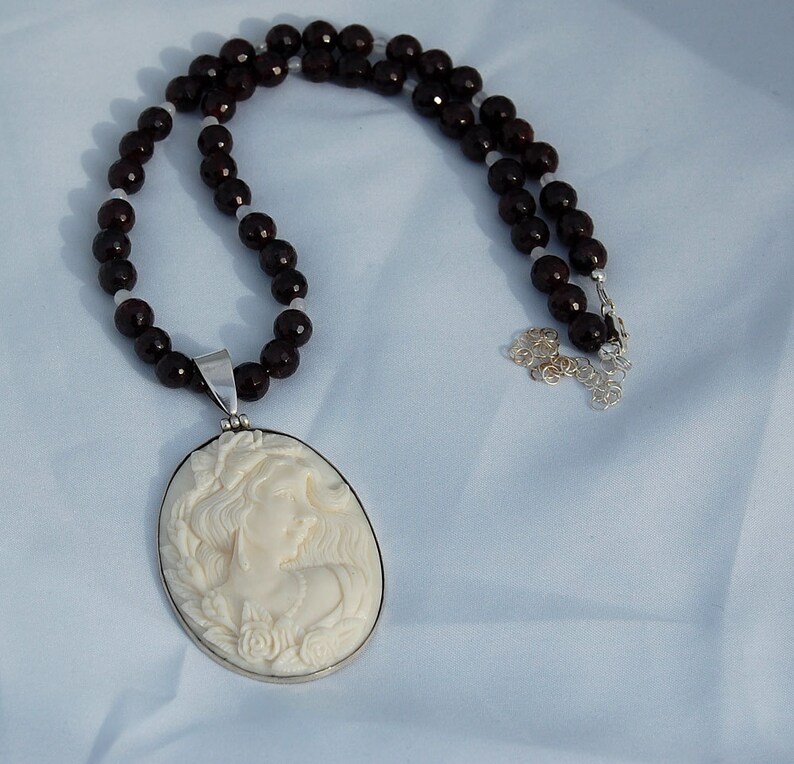 K&ouml;nnte beinhalten: Eine Halskette mit einem cremefarbenen Cameo-Anh&auml;nger, der das Profil einer Frau zeigt, in einem silbernen Rahmen. Die Halskette hat eine Perlenkette aus dunkelroten und wei&szlig;en Perlen. Der Schmuck wird auf einem hellblauen Stoff pr&auml;sentiert.