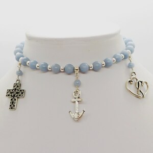 May include: A light blue beaded choker necklace with silver-toned charms. The charms include a cross, an anchor, and a double heart. The necklace is displayed on a white mannequin bust.