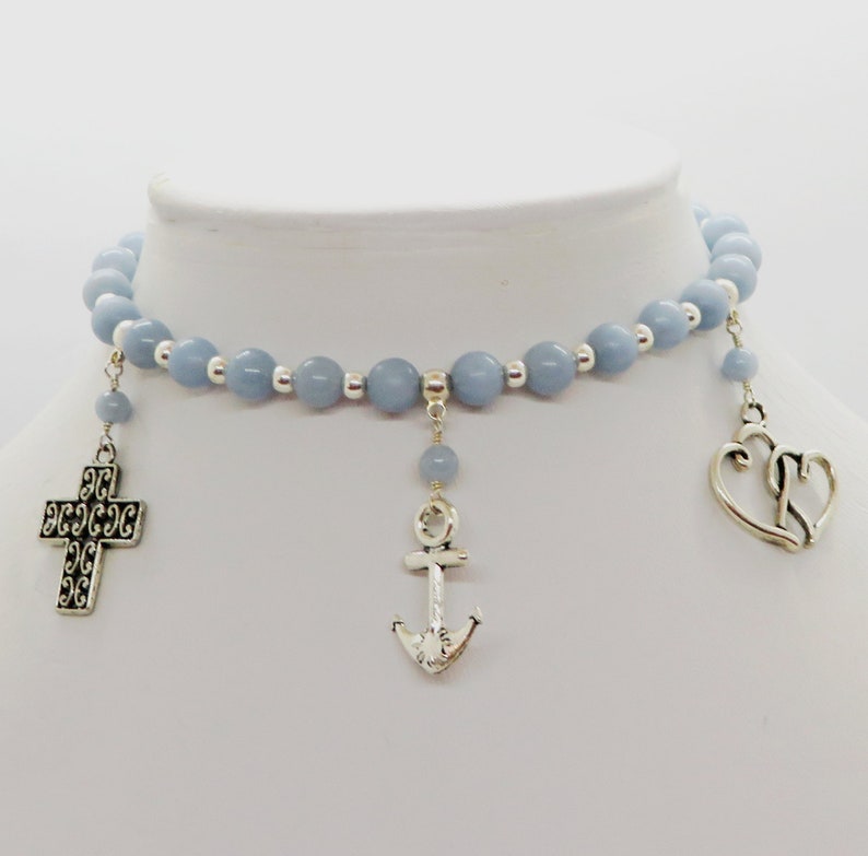 May include: A light blue beaded choker necklace with silver-toned charms. The charms include a cross, an anchor, and a double heart. The necklace is displayed on a white mannequin bust.