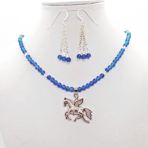 May include: A blue beaded necklace and matching earrings set. The necklace features a silver pendant of a winged horse. The earrings have a silver chain with blue and clear beads. The jewelry is displayed on a white mannequin.