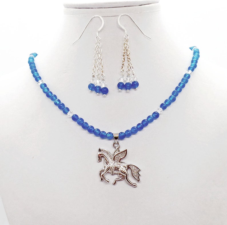 May include: A blue beaded necklace and matching earrings set. The necklace features a silver pendant of a winged horse. The earrings have a silver chain with blue and clear beads. The jewelry is displayed on a white mannequin.