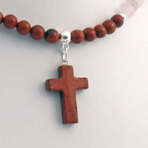 May include: A necklace featuring a reddish-brown stone cross pendant. The cross hangs from a silver-colored bail attached to a strand of reddish-brown and pink beads. The beads are round and evenly spaced.