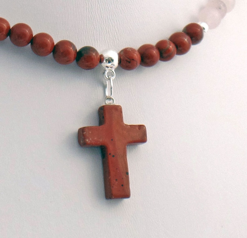 May include: A necklace featuring a reddish-brown stone cross pendant. The cross hangs from a silver-colored bail attached to a strand of reddish-brown and pink beads. The beads are round and evenly spaced.
