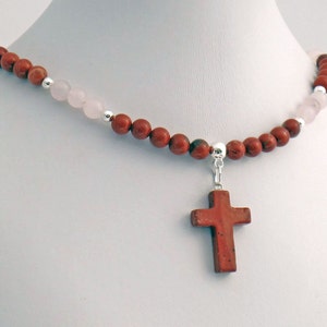 May include: A necklace featuring a cross pendant. The necklace is composed of red and pink beads, with silver accents. The cross pendant is a reddish-brown color. The necklace is displayed on a white mannequin.
