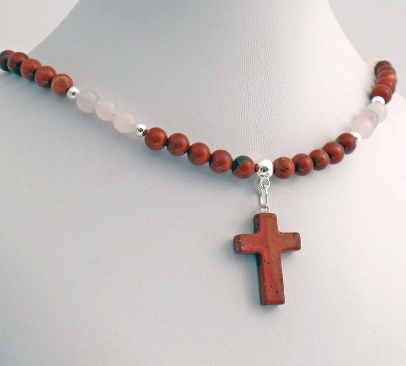 May include: A necklace featuring a cross pendant. The necklace is composed of red and pink beads, with silver accents. The cross pendant is a reddish-brown color. The necklace is displayed on a white mannequin.