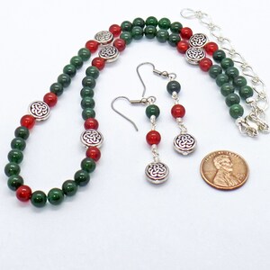 May include: A beaded jewelry set featuring a necklace and earrings. The necklace and earrings have green and red beads with silver-colored Celtic knot accents. The earrings hang from silver-colored hooks. A penny is included for size reference.