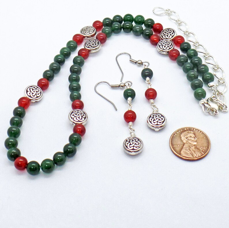 May include: A beaded jewelry set featuring a necklace and earrings. The necklace and earrings have green and red beads with silver-colored Celtic knot accents. The earrings hang from silver-colored hooks. A penny is included for size reference.