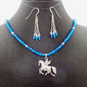 May include: A jewelry set featuring a blue beaded necklace with a silver Pegasus pendant, and matching dangle earrings. The earrings have silver chains and blue and clear crystal accents. The necklace and earrings are displayed on a black mannequin.