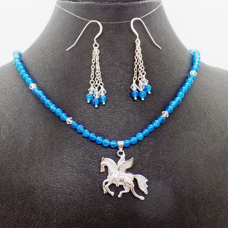 May include: A jewelry set featuring a blue beaded necklace with a silver Pegasus pendant, and matching dangle earrings. The earrings have silver chains and blue and clear crystal accents. The necklace and earrings are displayed on a black mannequin.