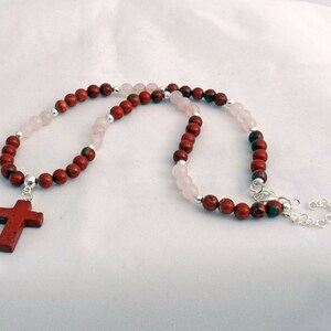 May include: A necklace featuring a red cross pendant and alternating red and pink beads. The necklace has a silver chain and clasp. The beads are strung on a silver chain. The cross is a darker red color.