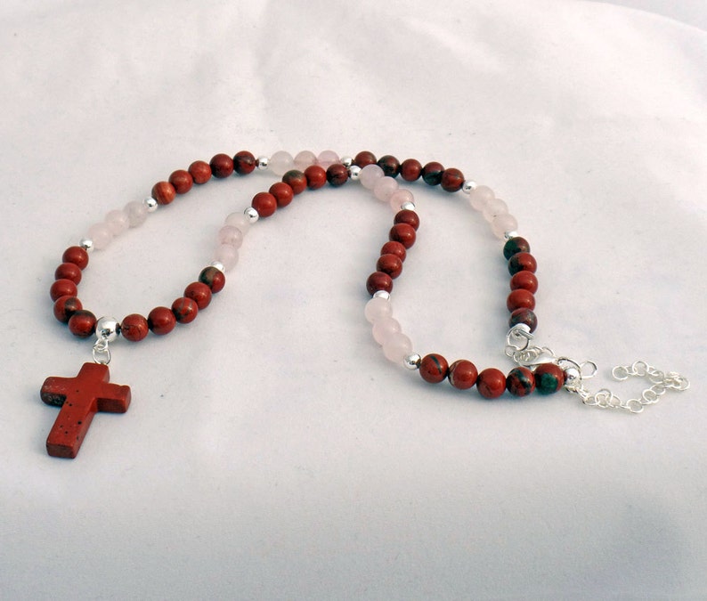 May include: A necklace featuring a red cross pendant and alternating red and pink beads. The necklace has a silver chain and clasp. The beads are strung on a silver chain. The cross is a darker red color.
