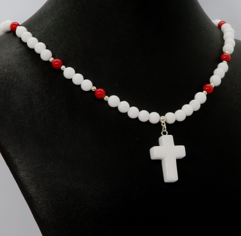 May include: White beaded necklace with a white cross pendant. The necklace features alternating white and red beads, with a silver-toned clasp. The cross pendant is a solid white color.