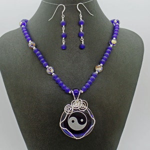 May include: A necklace and earrings set featuring a yin yang symbol pendant. The necklace is made with dark blue glass beads and the pendant is surrounded by silver wire. The earrings are silver with dark blue glass beads.