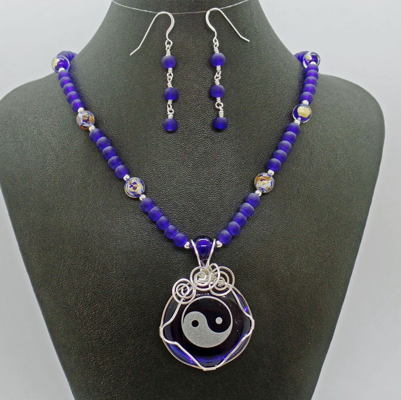 May include: A necklace and earrings set featuring a yin yang symbol pendant. The necklace is made with dark blue glass beads and the pendant is surrounded by silver wire. The earrings are silver with dark blue glass beads.