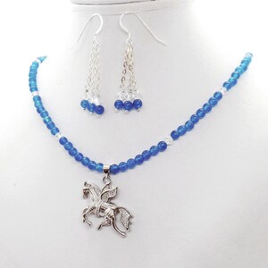 May include: A jewelry set featuring a blue beaded necklace with a silver Pegasus pendant, and matching dangle earrings. The earrings and necklace have clear and blue beads. The Pegasus pendant is detailed with clear stones.