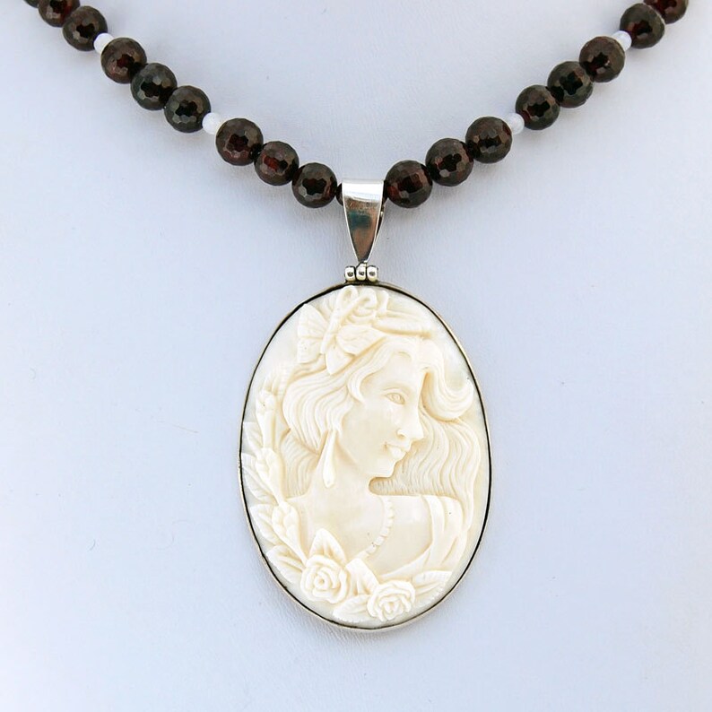 K&ouml;nnte beinhalten: Eine cremefarbene Cameo-Anh&auml;nger-Halskette mit dem Profil einer Frau, silberfarben gerahmt und an einer Perlenkette aus dunkelroten und wei&szlig;en Perlen aufgereiht. Das Cameo zeigt eine Frau mit flie&szlig;endem Haar und floralen Akzenten.