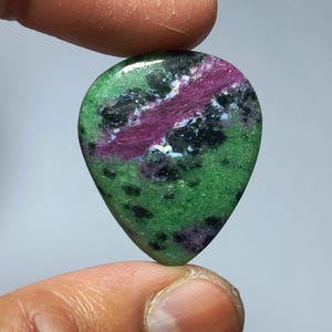 Puede incluir: Una gema en forma de lágrima con una vibrante mezcla de colores. La piedra presenta una base verde oscuro salpicada de puntos negros y una prominente banda de magenta intenso. Acentos blancos añaden contraste.