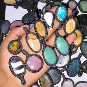 Peut inclure: Une collection de pendentifs en pierres précieuses de différentes formes et couleurs, dont ovales, en forme de goutte et ronds. Chaque pierre est sertie dans un cadre en macramé noir. Les pierres précieuses affichent des couleurs variées.