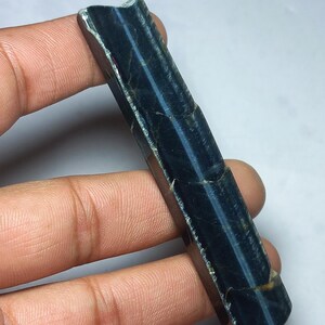May include: A rectangular gemstone with a polished surface, displaying a deep blue colour with light blue stripes. The stone is held in a hand, highlighting its natural texture and unique pattern. The gemstone appears to be a Tiger's Eye variant.