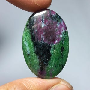 Op de afbeelding: Een ovale edelsteen met een gepolijst oppervlak. De steen heeft een mix van kleuren, waaronder diepgroen, zwart en een levendig magenta. De edelsteen wordt tussen twee vingers gehouden, wat de grootte en vorm laat zien.