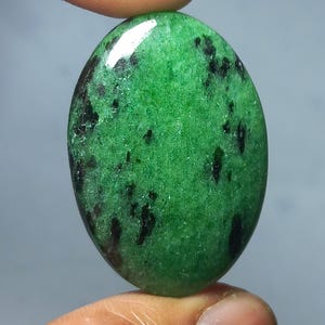 Könnte beinhalten: Oval geformter grüner Edelstein mit schwarzen Einschlüssen. Der polierte Stein hat eine glänzende Oberfläche und wird zwischen zwei Fingern gehalten. Der Edelstein hat eine leuchtend grüne Farbe mit dunklen Flecken.