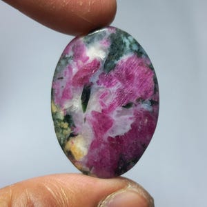Puede incluir: Una gema ovalada con una mezcla de colores. La piedra presenta un vibrante tono rosa con parches blancos, verdes y amarillos. La gema pulida se sostiene entre dos dedos, mostrando sus patrones naturales y su textura.