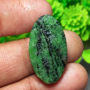 May include: An oval-shaped, rough-cut gemstone with a vibrant green base color and striking black inclusions. The stone is held in a hand, showcasing its natural texture and unique pattern. The gemstone's surface appears slightly uneven, highlighting its raw, unpolished state.