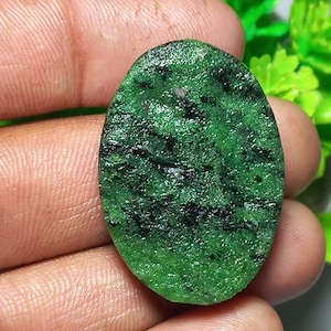 May include: Oval-shaped green gemstone with black inclusions. The polished stone has a textured surface and is held in a hand. The gemstone is set against a blurred background of green foliage.