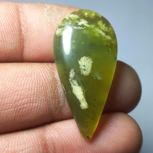 Può includere: Una gemma a forma di goccia lucidata. La pietra presenta una miscela di tonalità verde oliva e giallo, con inclusioni bianco crema. La gemma è tenuta in mano, evidenziando la sua forma e dimensione.