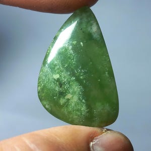 Può includere: Una gemma lucida a forma di goccia, tenuta tra le dita. La pietra è di un verde vibrante con motivi più chiari e screziati e una superficie lucida. La gemma ha una forma liscia e arrotondata, con una leggera punta nella parte superiore.