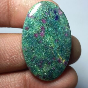 Peut inclure: Pierre précieuse ovale avec une base verte mouchetée de petites inclusions rouges et bleu foncé. La surface polie reflète la lumière, soulignant les textures et motifs naturels de la pierre. Une gemme naturelle unique.
