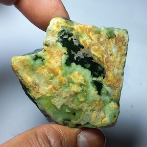 Può includere: Una gemma grezza con una miscela di tonalità verdi, gialle e marroni. La pietra ha una superficie strutturata e una forma irregolare, con aree di verde scuro e verde-giallo più chiaro. La gemma è tenuta dalle dita di una persona.