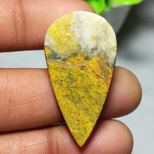 May include: A teardrop-shaped gemstone with a vibrant yellow and white color scheme. The stone features a textured surface with a mix of yellow, white, and hints of red and black. The gemstone is held in a hand.