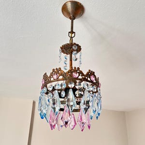 Puede incluir: Una lámpara de techo con una estructura circular de metal en tono bronce, adornada con colgantes de cristal facetado que alternan entre azul claro y rosa. La luminaria incluye una varilla central y un dosel decorativo.