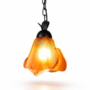 May include: An orange and yellow glass pendant light with a floral design. The light fixture is suspended from a black chain and has a black decorative top. The glass shade has a wavy, organic shape and is illuminated from within.