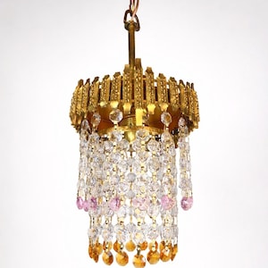 May include: A vintage chandelier with a gold-toned metal frame and a cascade of clear and pink crystal beads. The chandelier hangs from a chain and features amber-coloured crystal accents at the bottom. A decorative lighting fixture.