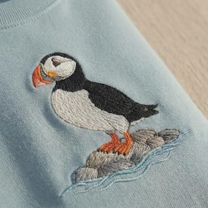 Pode incluir: Camisola azul clara com um bordado de um papagaio-do-mar. A ave, com a sua plumagem preta e branca e o bico e patas laranja, está pousada numa formação rochosa com água bordada. O design é detalhado.