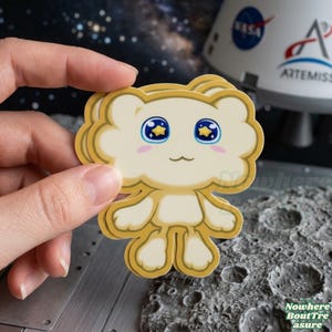 May include: A die-cut sticker featuring a cartoon character with a cloud-like head, light yellow body, and blue eyes with star details. The sticker is held in a hand against a space-themed background.
