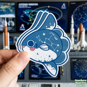 May include: A die-cut sticker featuring a blue and white sunfish design with a constellation pattern. The sticker has a cartoon-like appearance with a smiling face and is held in a hand. The background includes images of rockets and space-themed displays.