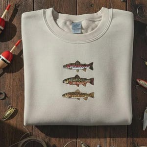 May include: A beige sweatshirt featuring embroidered illustrations of three different fish. Fishing bobbers and lures are scattered around the sweatshirt on a wooden surface, suggesting a fishing theme.
