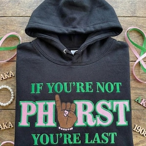 May include: Black hooded sweatshirt with green and pink embroidered text that reads "IF YOU'RE NOT PHIRST YOU'RE LAST". The design includes a raised hand with a pearl bracelet. The sweatshirt has a drawstring hood.